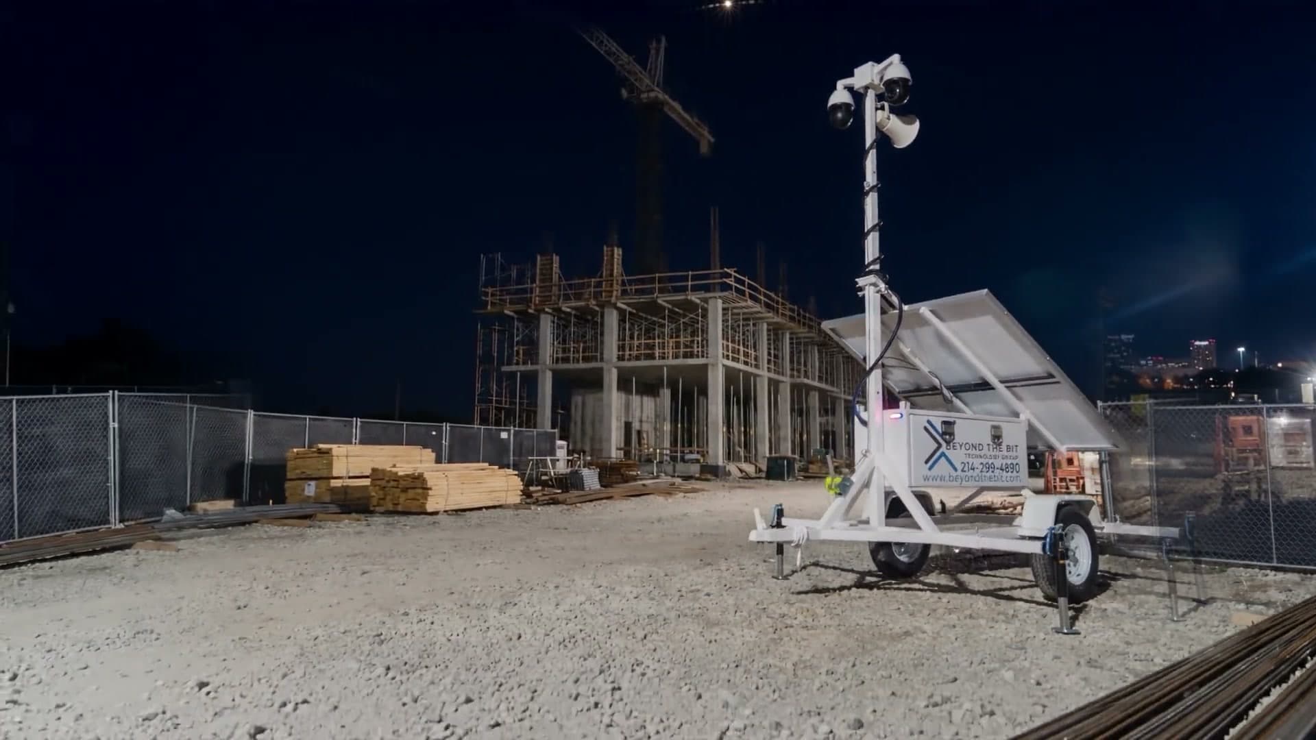 A Beyond the Bit security trailer on a commercial construction site at night, camera mast fully extended above a concrete-frame building under construction, tower crane and stacked lumber behind the perimeter fence.