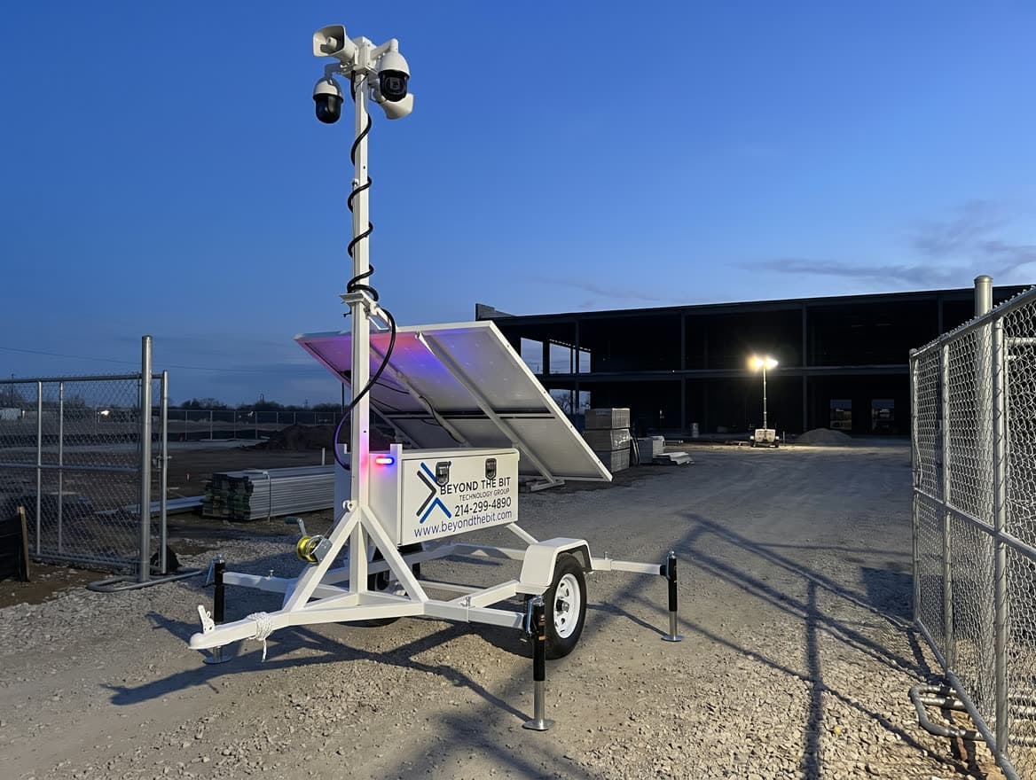A security trailer watching the vehicle gate of a Dallas–Fort Worth jobsite at dusk — the ground a guard would patrol on foot and a camera trailer watches continuously.