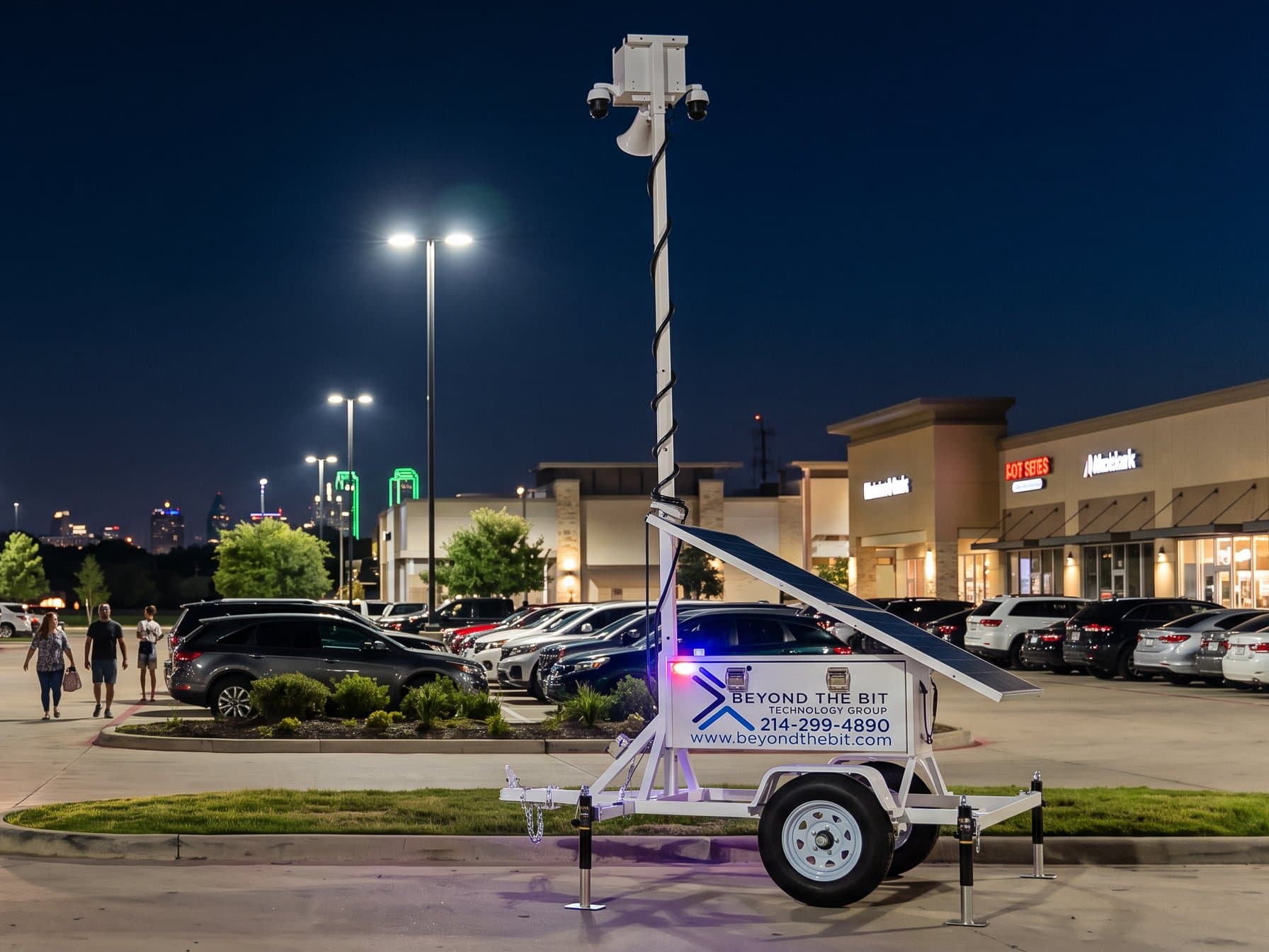 A Beyond the Bit security trailer with its mast raised at the edge of a busy Dallas-area retail plaza at night, storefronts lit and shoppers walking to their cars.