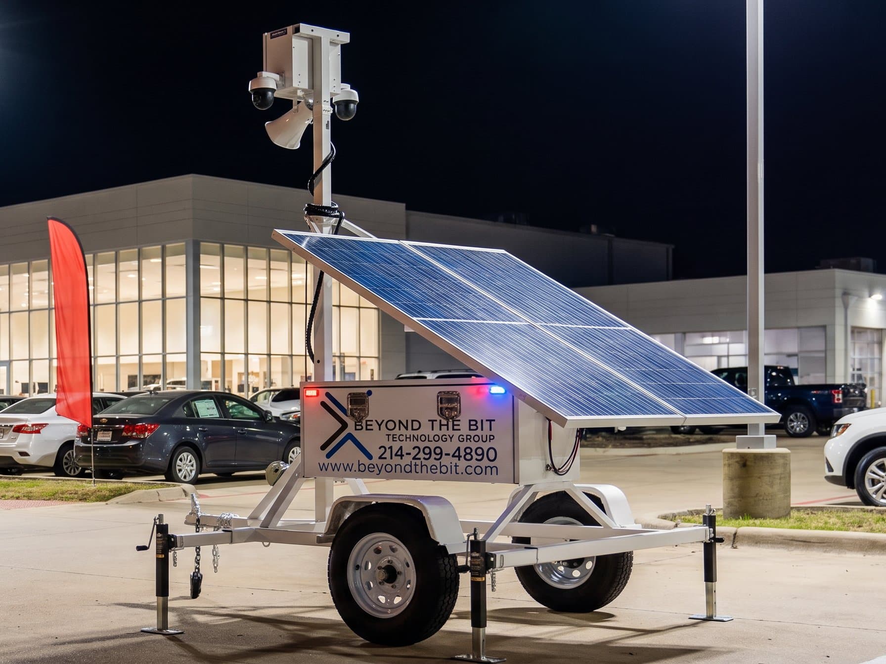 A Beyond the Bit security trailer with its mast raised at the corner of a new-car dealership lot at night, showroom glass glowing behind rows of inventory.