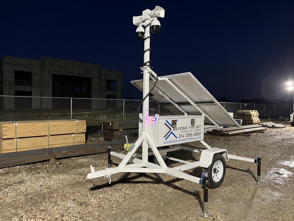 A Beyond the Bit security trailer standing watch at night in an equipment laydown yard, red-and-blue strobe bar lit, excavators and stacked pipe behind it.