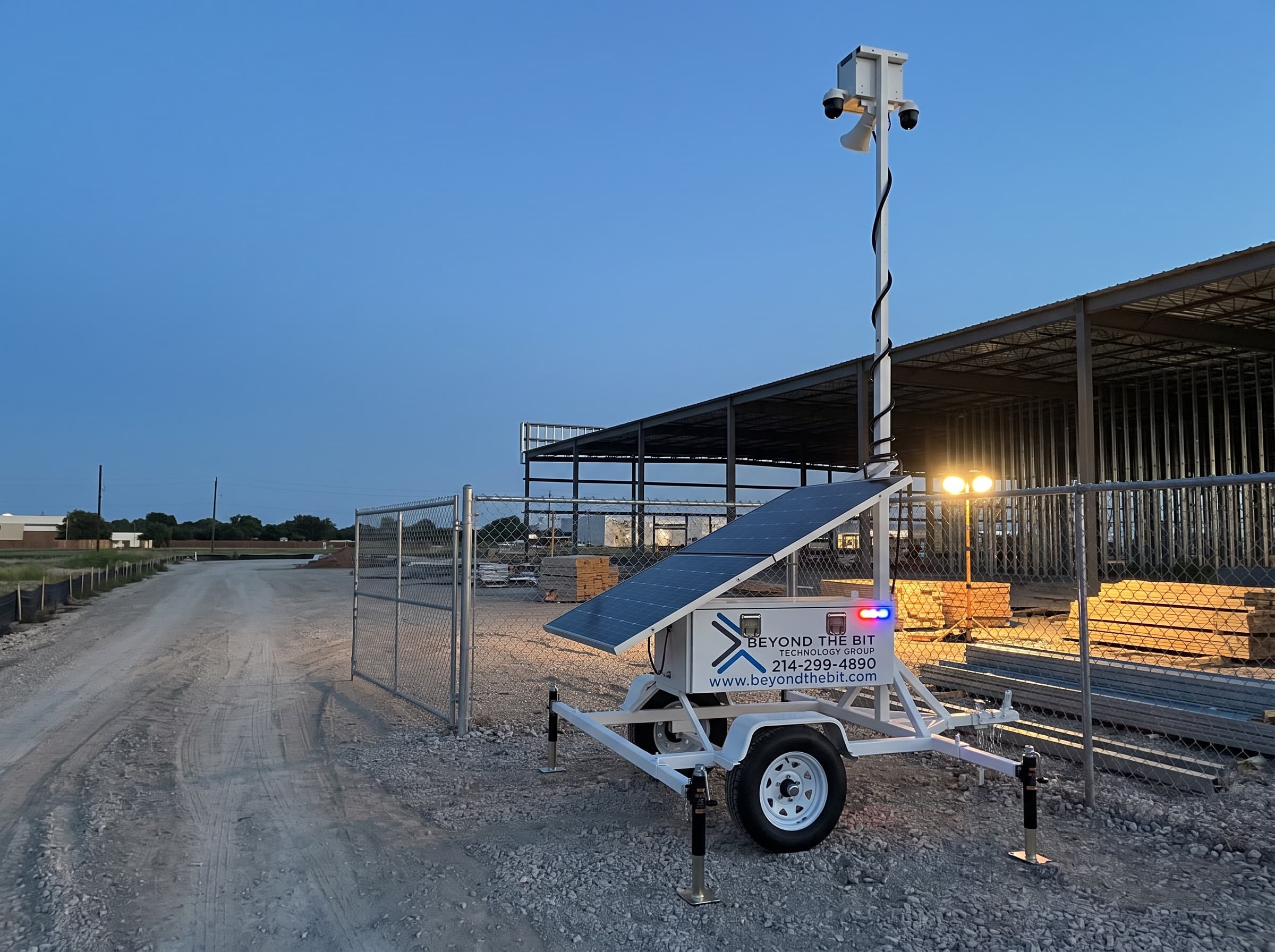 A Beyond the Bit security trailer parked on the shoulder just inside a fenced construction site entrance at dusk, camera mast raised, the open vehicle gate and drive lane clear beside it.